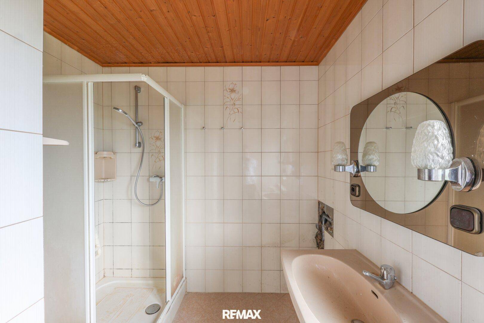 Badezimmer mit Duschkabine, Waschbecken und Holzdecke im 80er-Jahre-Stil, renovierungsbedürftig.
