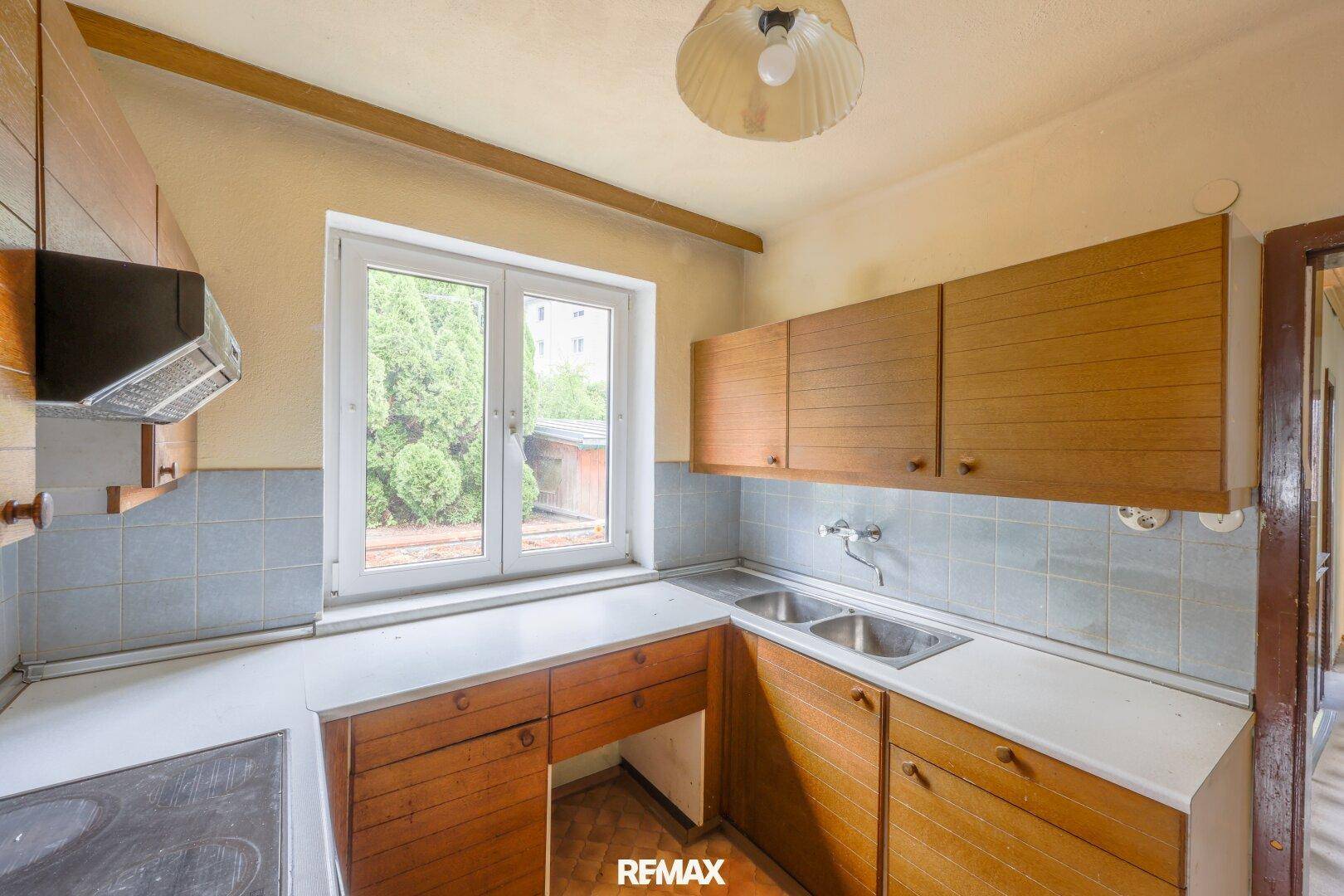 Küche mit Holzschränken, Doppelspüle und Fensterblick ins Grüne, benötigt Modernisierung.