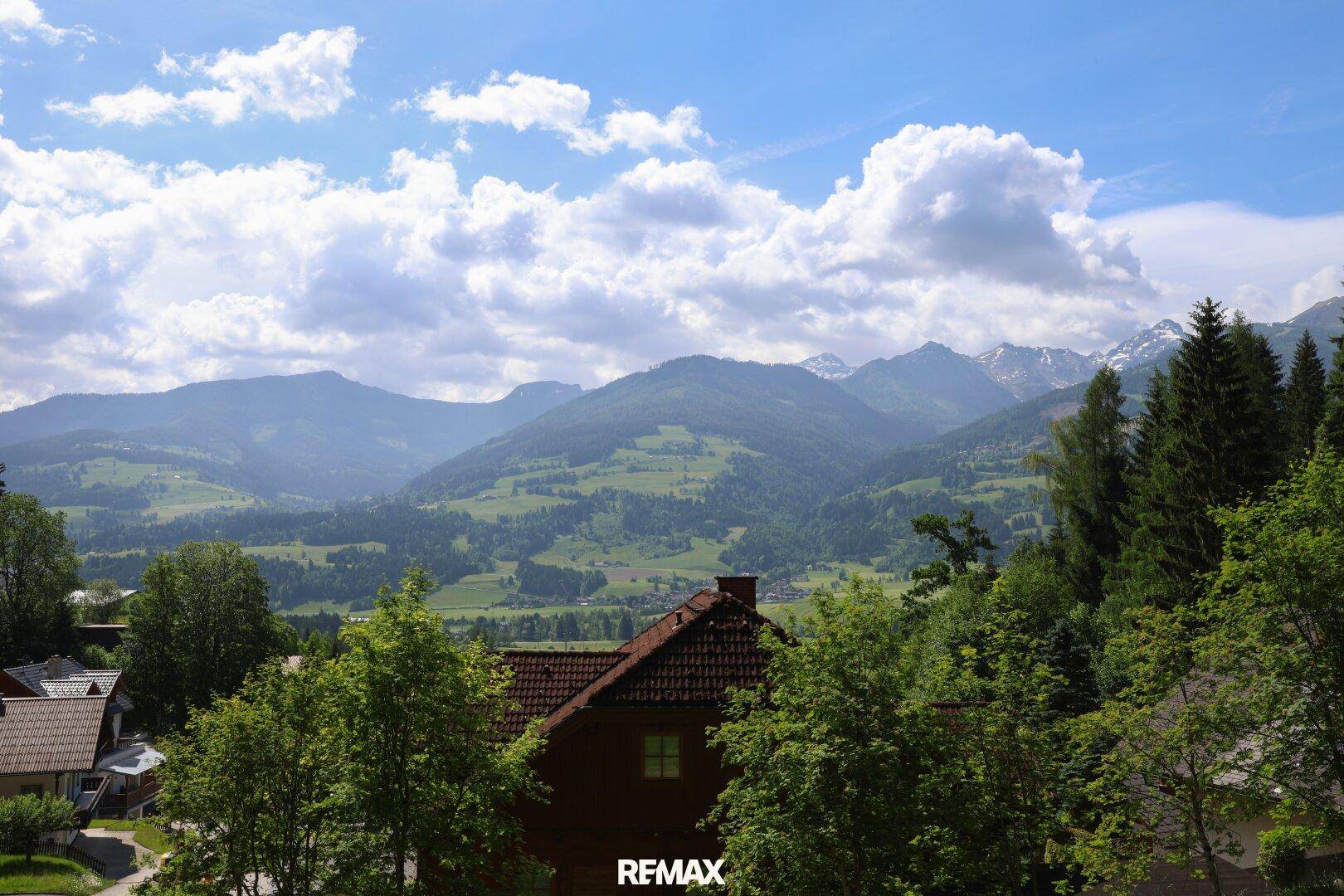 Panoramablick auf die umliegende Berglandschaft mit grünen Hügeln und vereinzelten Häusern unter blauem Himmel.