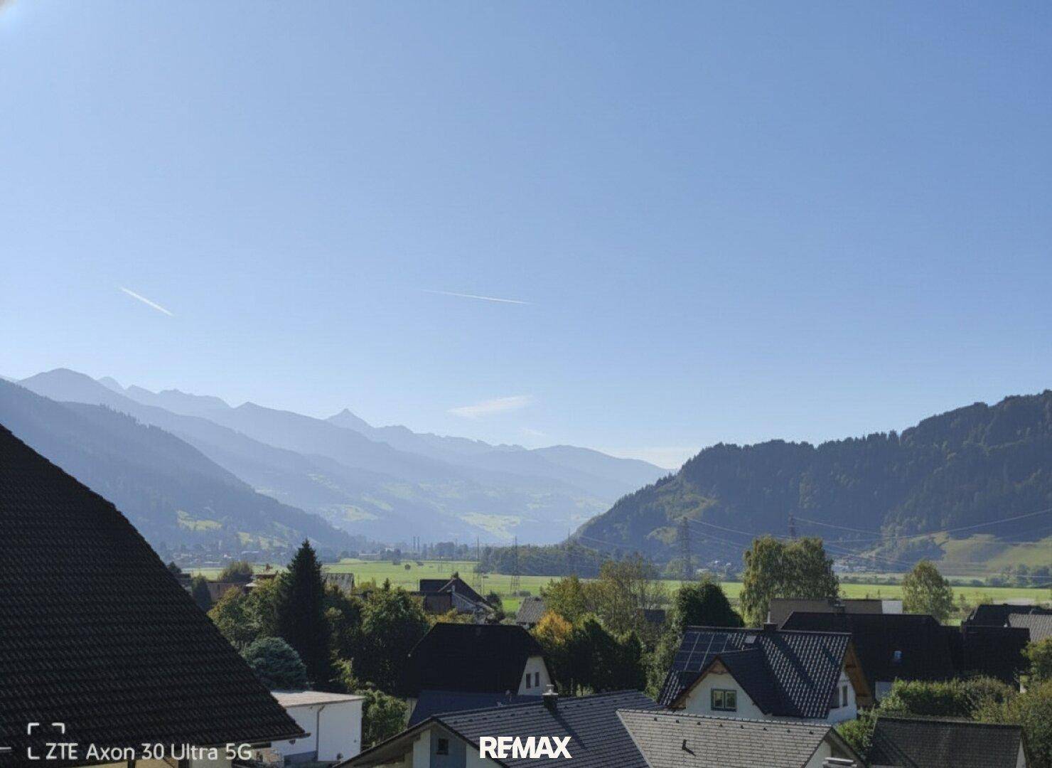 Weitläufiger Blick über ein Tal mit Siedlungen und majestätischen Bergen unter einem klaren blauen Himmel.