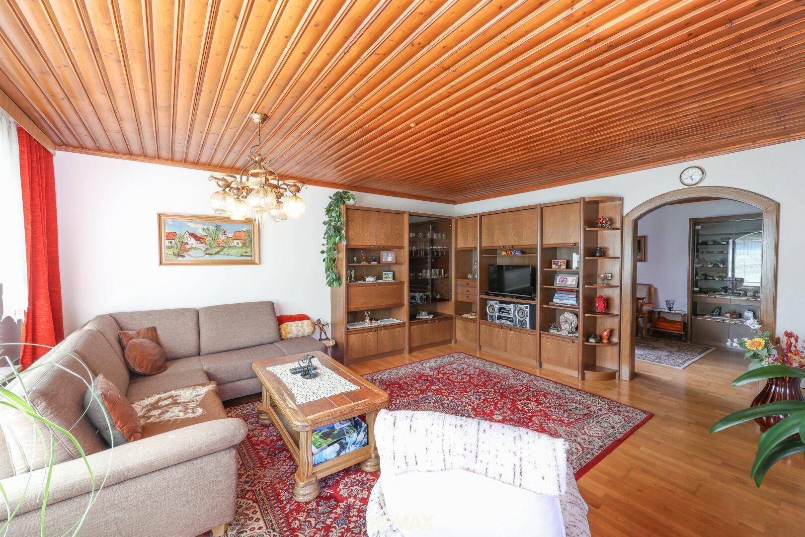 Wohnzimmer mit Holzdecke, umfangreicher Holzschrankwand und Zugang zu einem weiteren Raum.