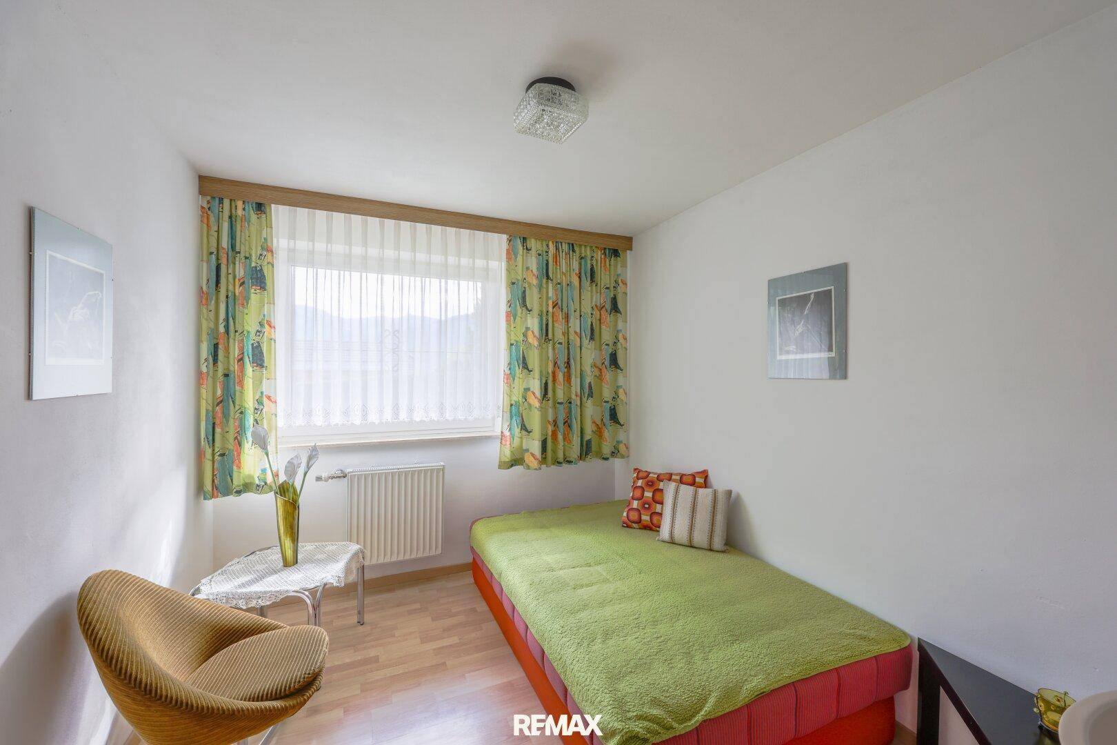 Einzelnes Schlafzimmer mit Bett, Stuhl und farbenfrohen Vorhängen, ideal als Kinderzimmer.