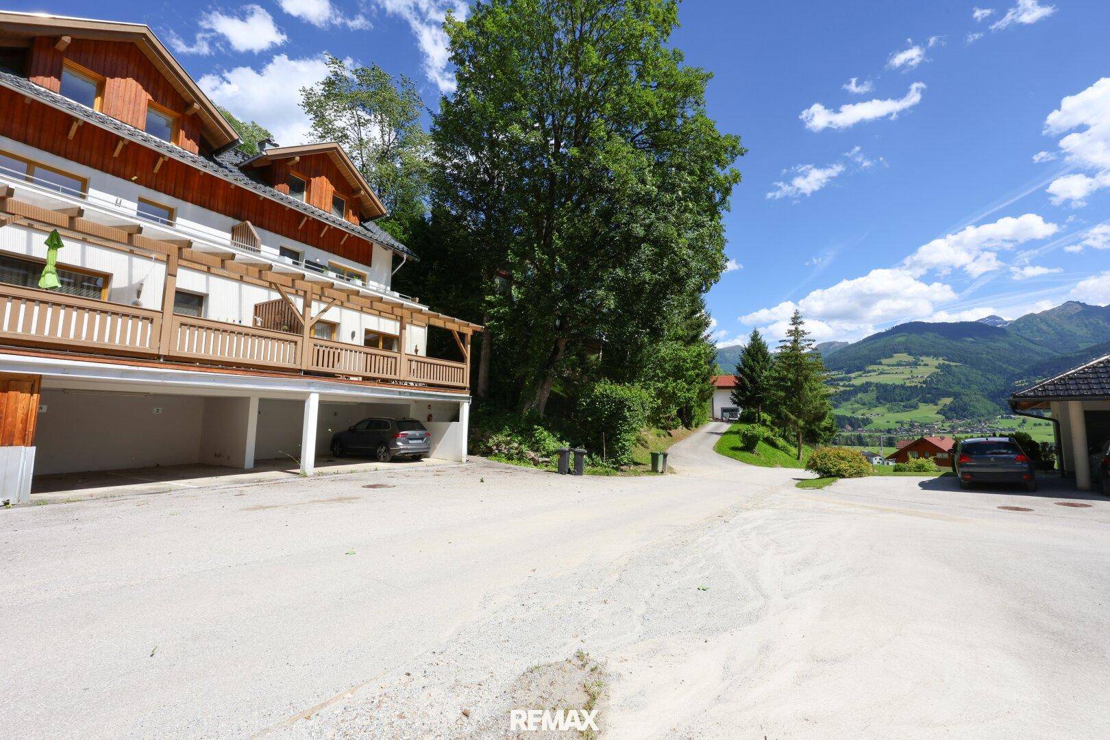 Traditionelles Gebäude im alpinen Stil mit Holzfassade, Balkonen und überdachten Parkplätzen, umgeben von grünen Bergen.