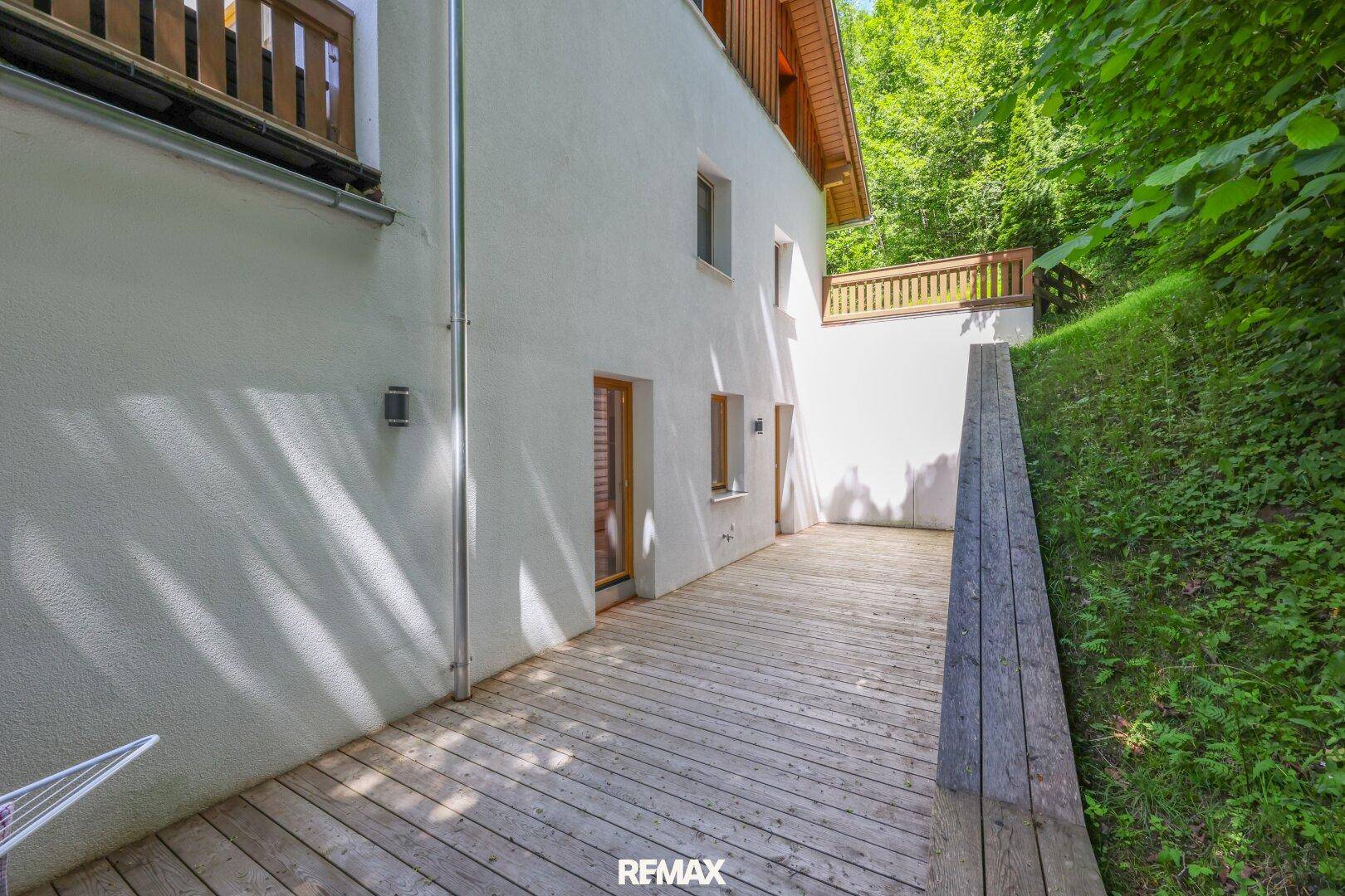 Holzterrasse am Hang des Gebäudes, umgeben von üppigem Grün und Bäumen.