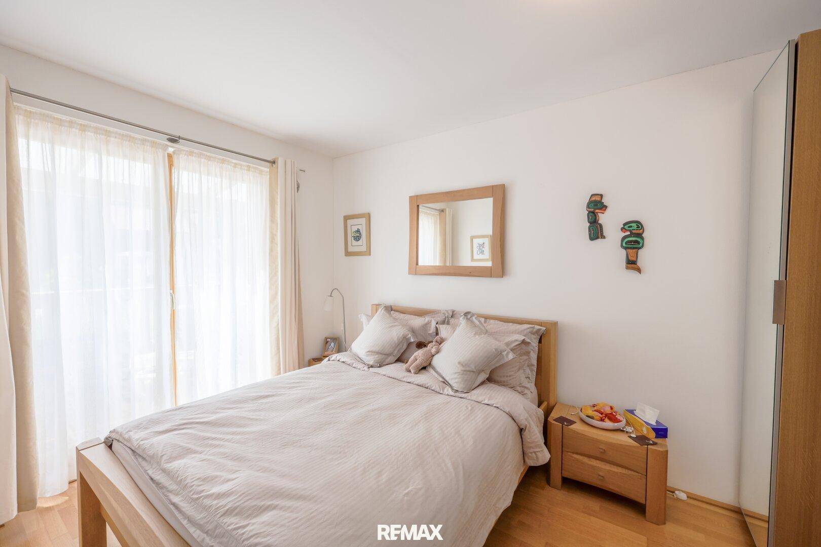 Helles Schlafzimmer mit Doppelbett, Holzmöbeln und großem Fenster mit Vorhängen.