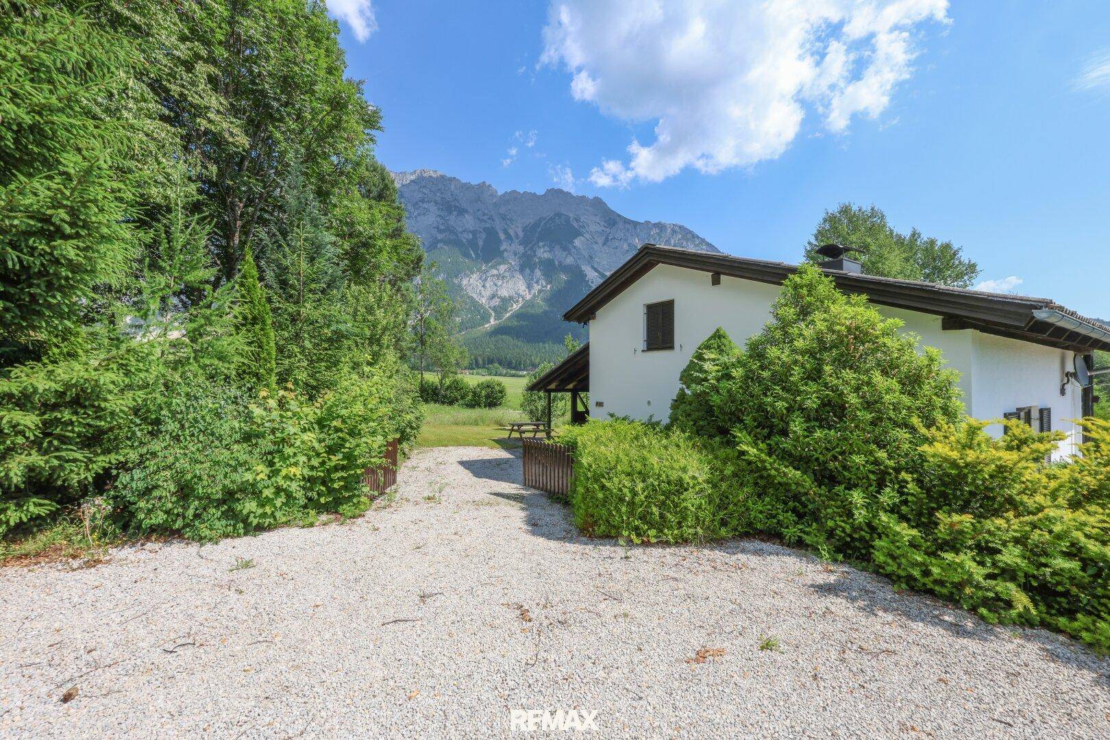 Zufahrt zum Haus mit Kiesweg, umgeben von Bäumen und Sträuchern, mit Bergblick.