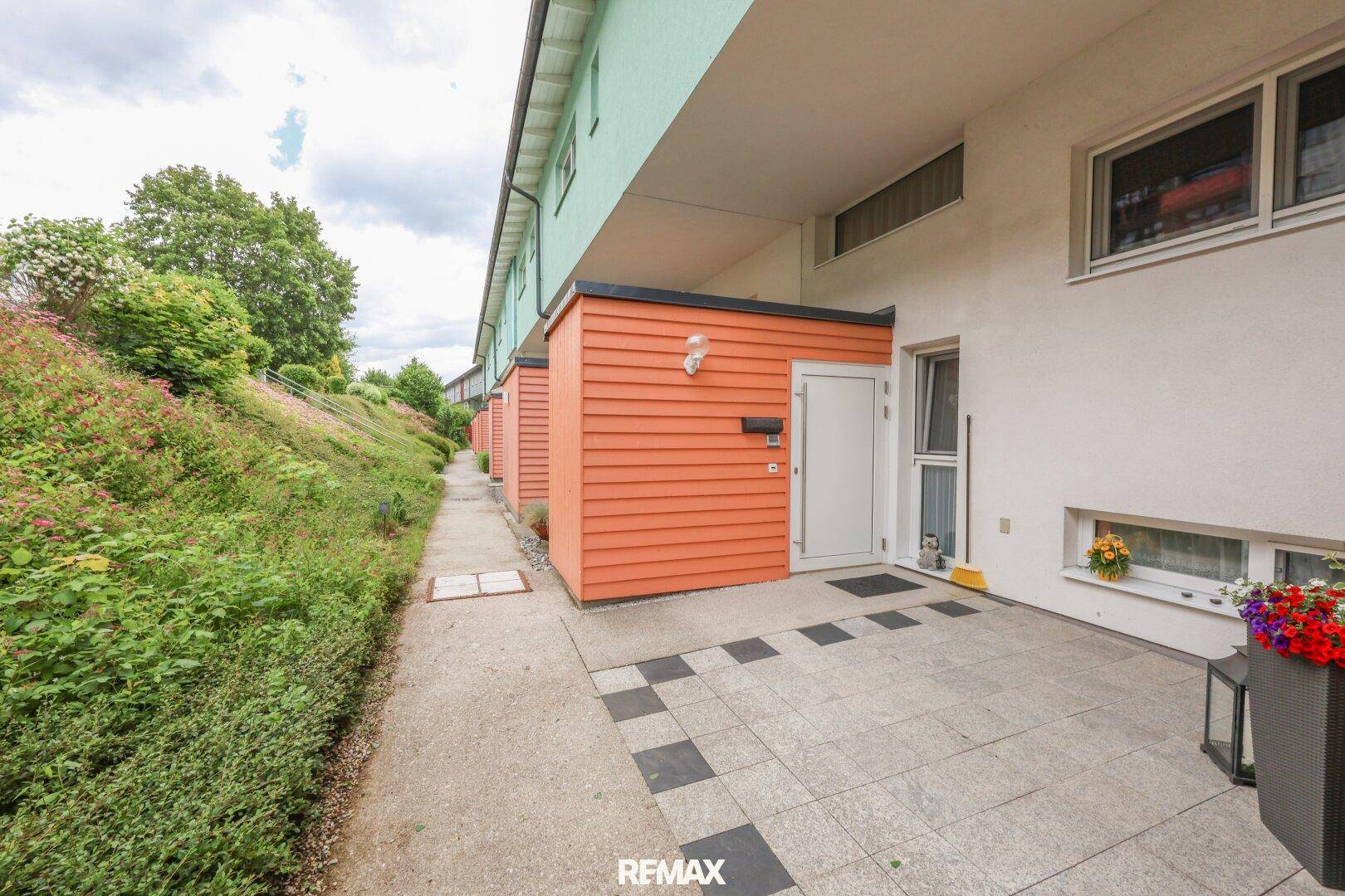 Eingangsbereich eines Reihenhauses mit kleiner gepflasterter Fläche und orangefarbener Fassadenverkleidung.
