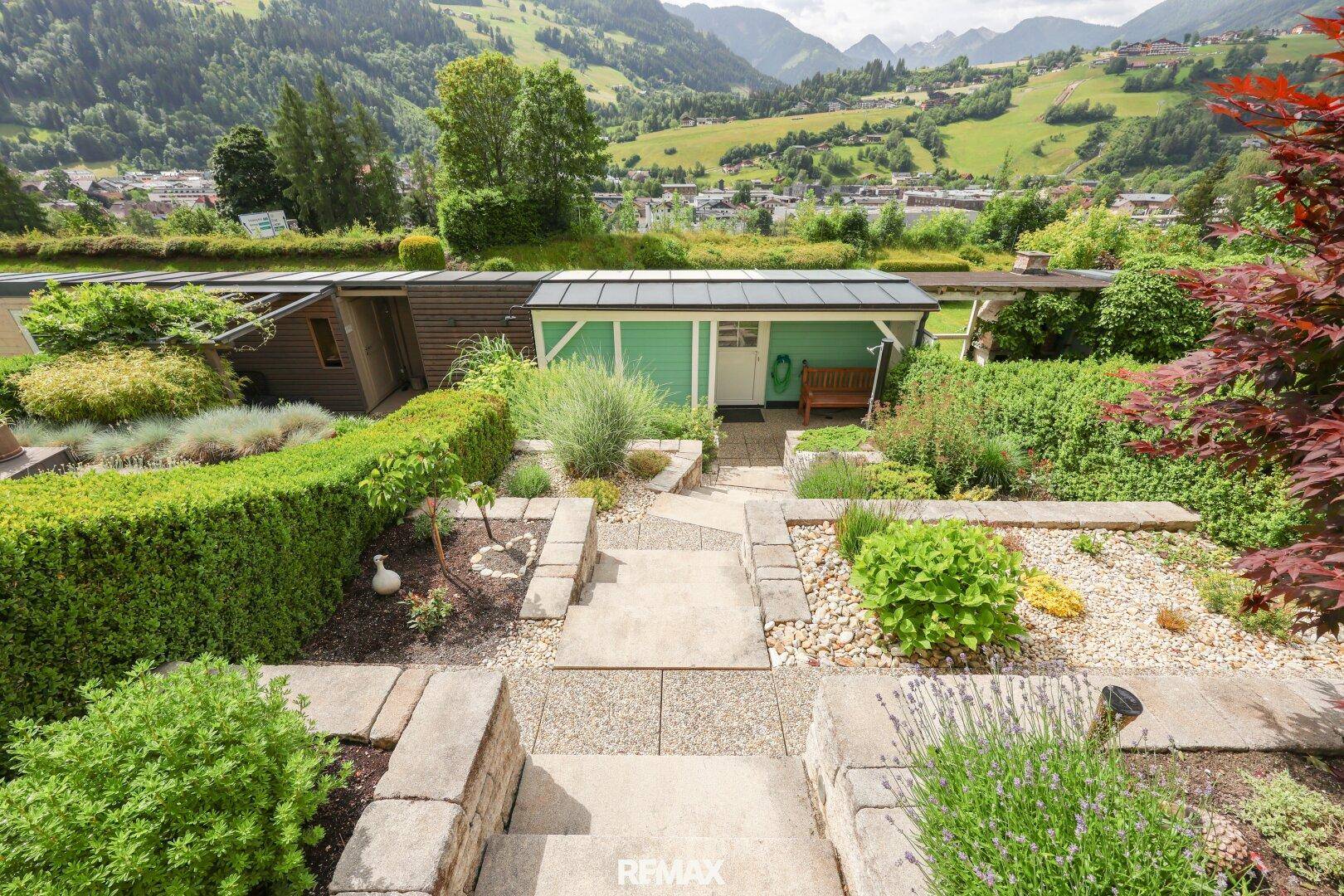 Gepflegter Garten mit Steinwegen, Hecken und einem kleinen grünen Gartenhaus vor alpiner Kulisse.