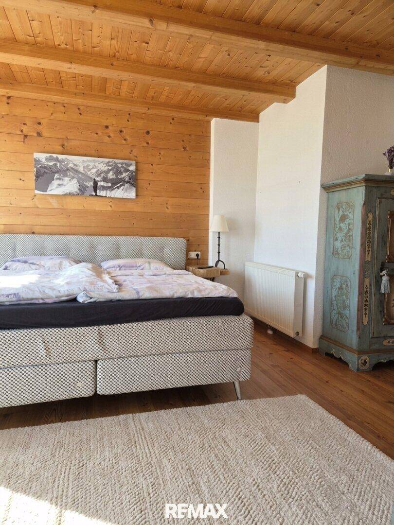 Helles Schlafzimmer mit Holzwänden, Holzbalkendecke, großem Bett und einem bemalten Bauernschrank.