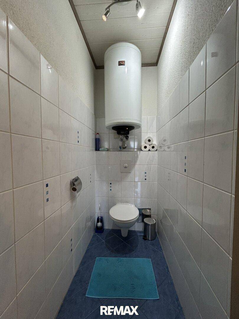 Kleines WC mit weißen Fliesen, blauem Boden und Warmwasserboiler.