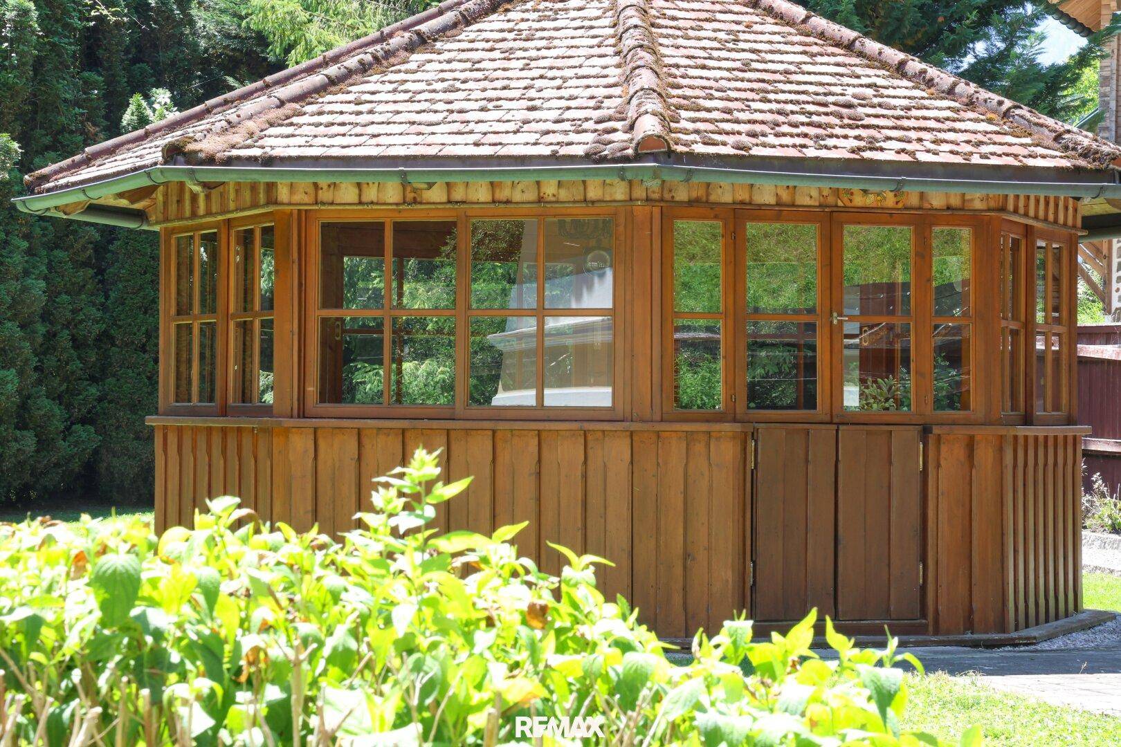 Holzpavillon im Garten mit vielen Fenstern und einem Ziegeldach.