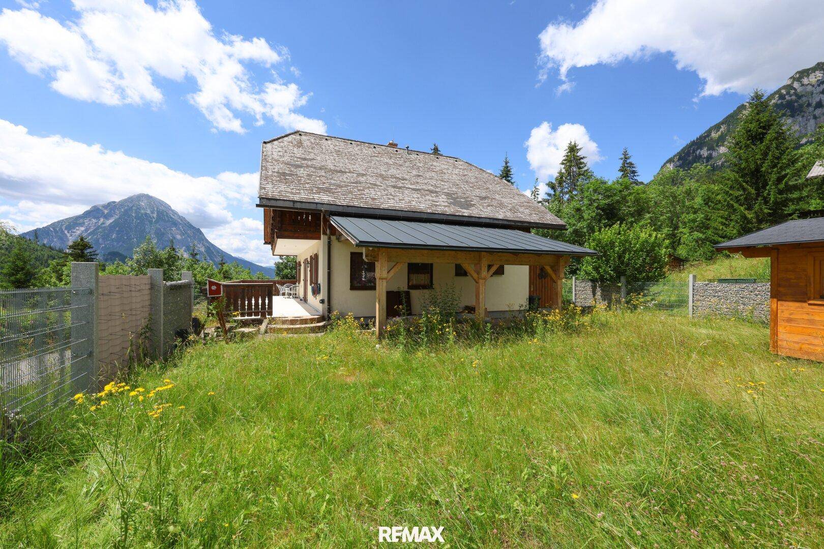 Charmantes Haus mit Holzveranda und weitläufigem Gartenbereich, umgeben von grüner Natur und Bergen.