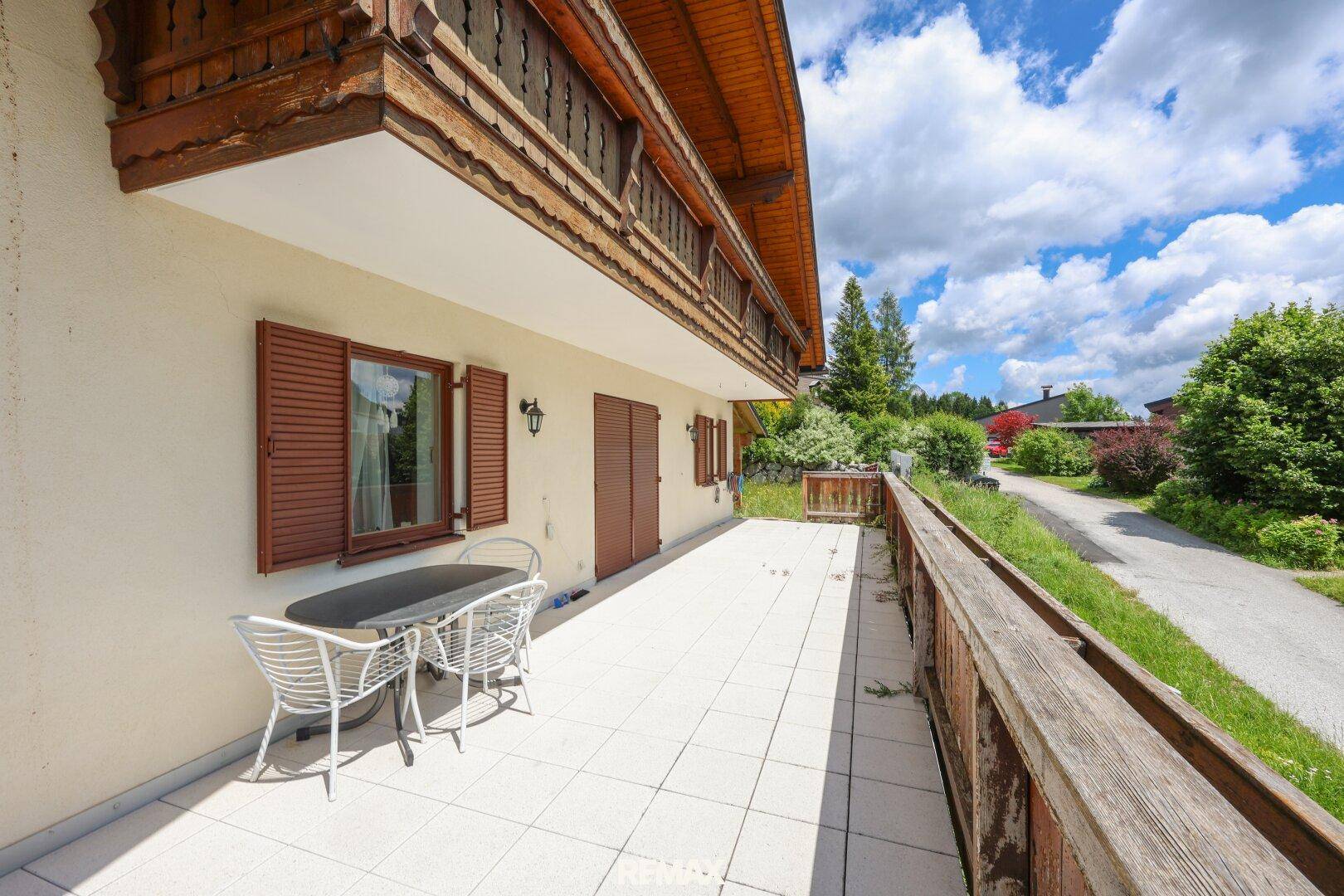 Helle Terrasse mit Fliesenboden und Gartenmöbeln, umgeben von grüner Landschaft und einem Weg.