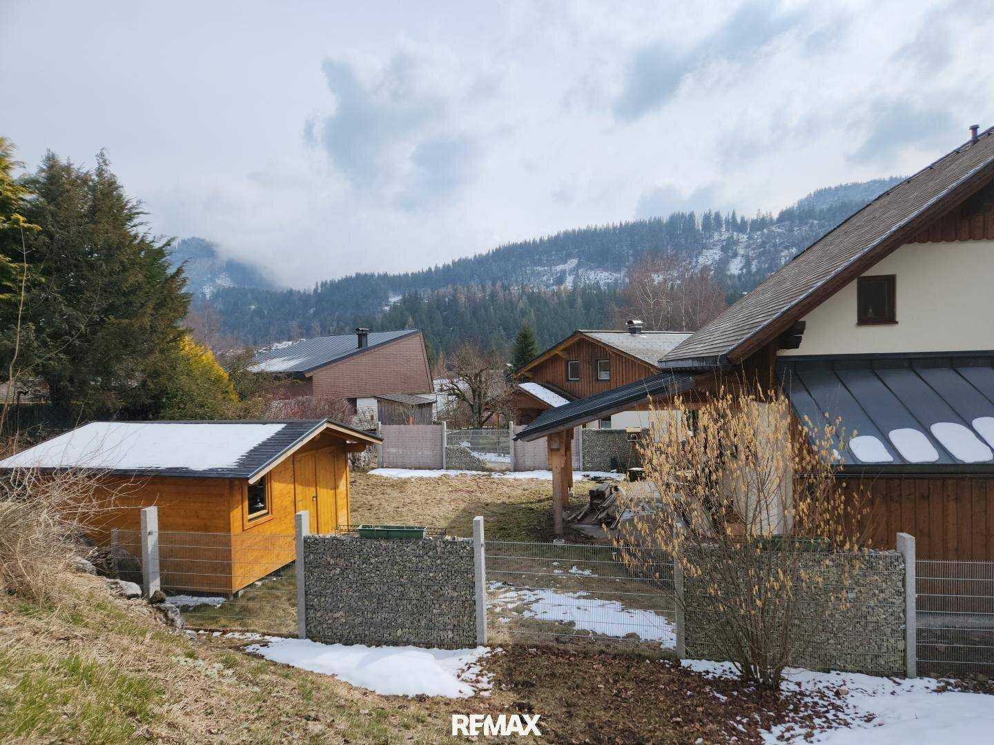 Winterliche Ansicht des Hauses mit Gartenbereich, umgeben von verschneiten Bergen und Bäumen.