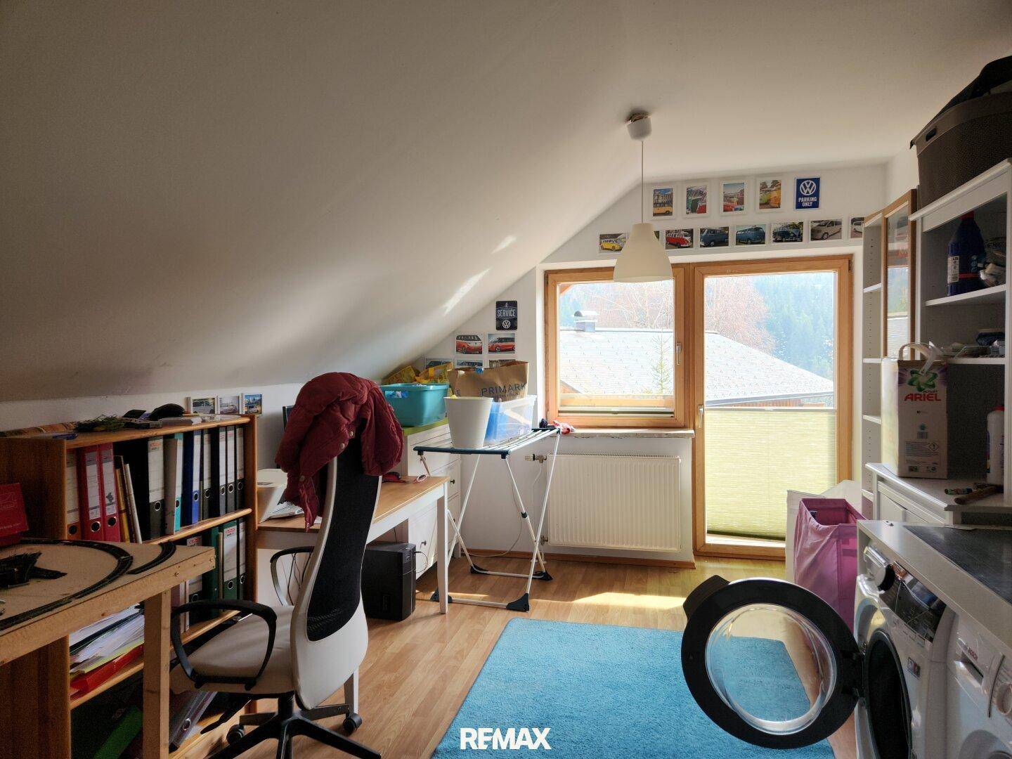 Dachgeschosszimmer mit Schreibtisch, Regalen und einer Waschmaschine, hell durch große Fenster.