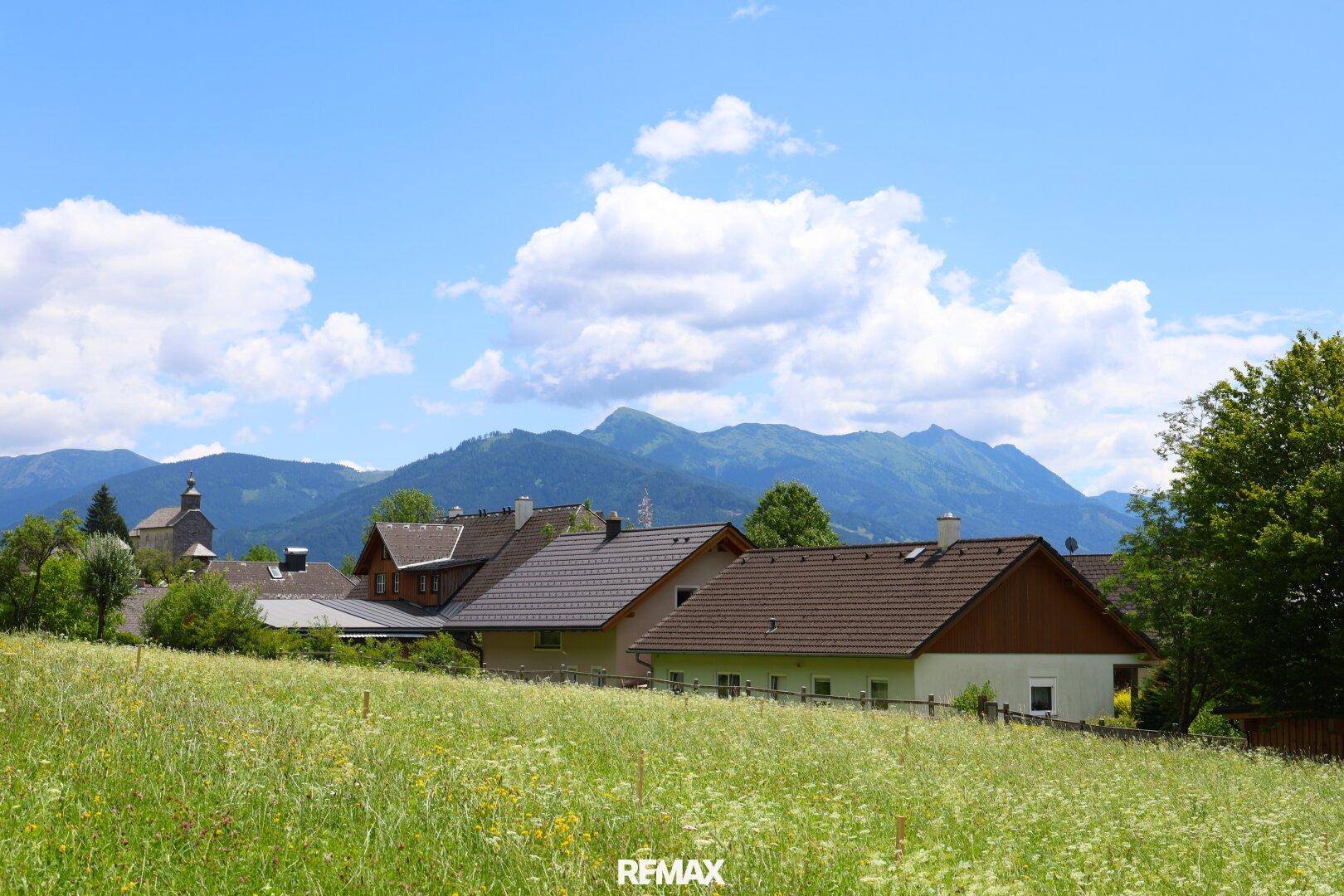Sonnige Landschaft mit Wohnhäusern, blühenden Wiesen und einer Bergkette im Hintergrund unter blauem Himmel.