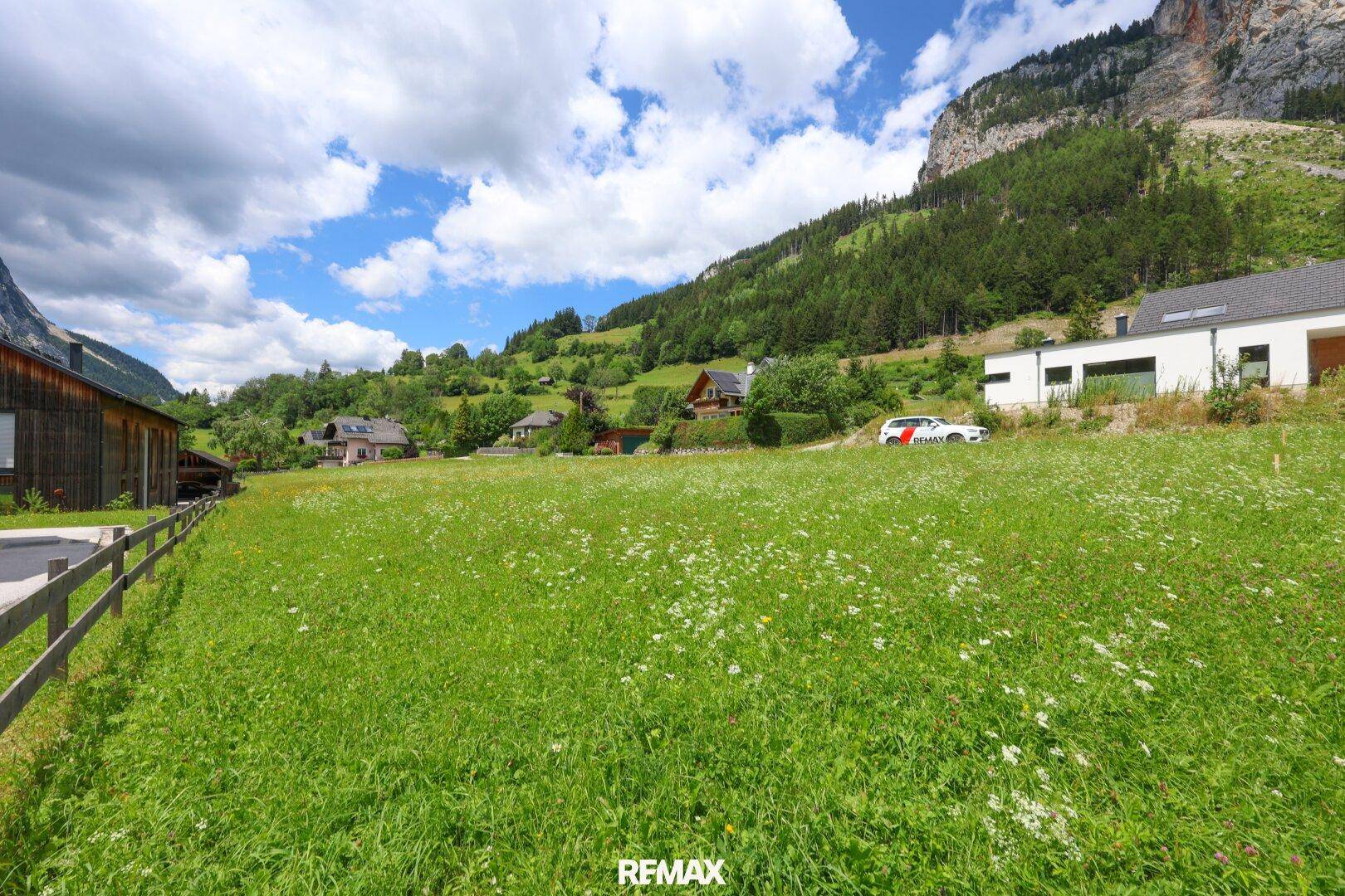 Weitläufige grüne Wiese mit Blick auf moderne Häuser und bewaldete Berghänge unter blauem Himmel.
