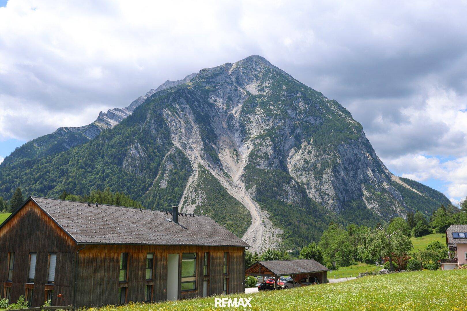 Modernes Holzhaus mit dunklem Dach vor einem imposanten Bergpanorama unter bewölktem Himmel.