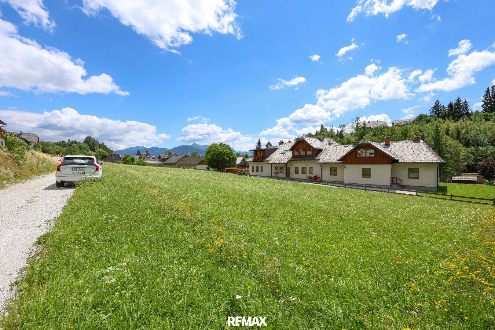 Idyllische Landschaft mit traditionellen Häusern, grünen Wiesen und Bergen unter strahlend blauem Himmel.