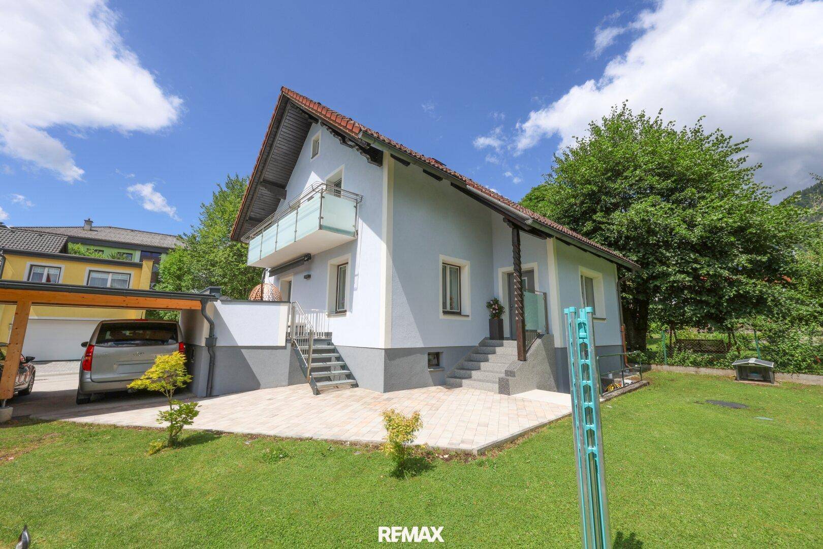 Freistehendes Haus mit hellblauer Fassade, gepflegtem Garten und Carport unter blauem Himmel.