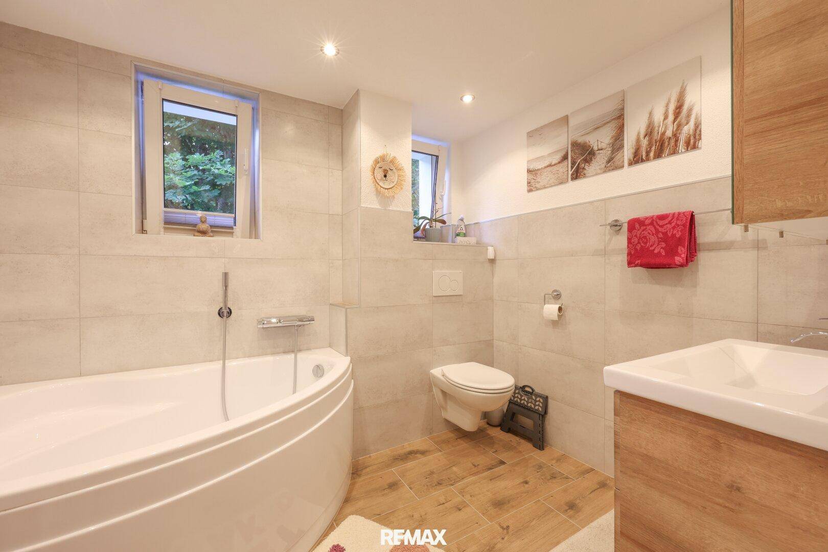 Modernes Badezimmer mit Eckbadewanne, Wand-WC, Waschbecken und zwei Fenstern.