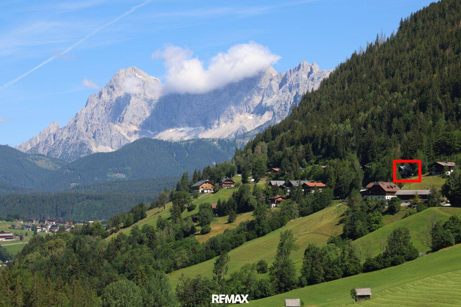 Malerische Berglandschaft mit traditionellen Häusern und einem markierten Objekt vor imposanten Gipfeln.