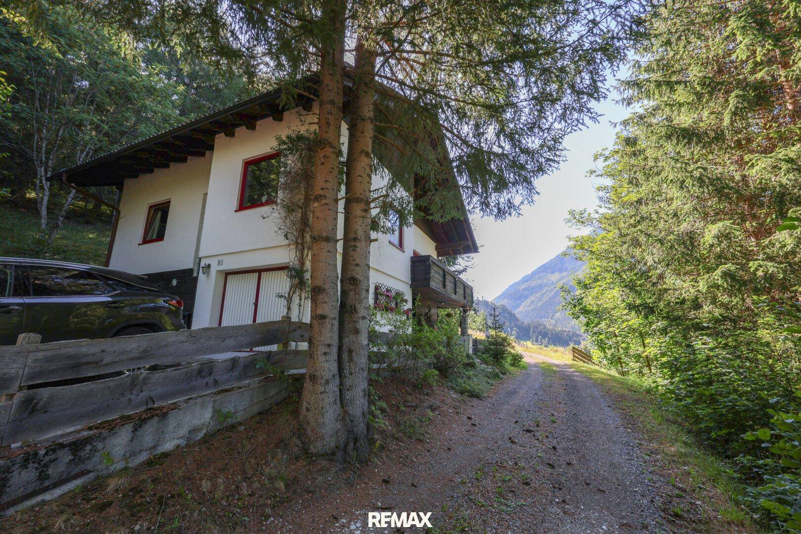 Charmantes Landhaus mit Holzbalkonen und einer Zufahrt, eingebettet in eine bewaldete Berglandschaft.