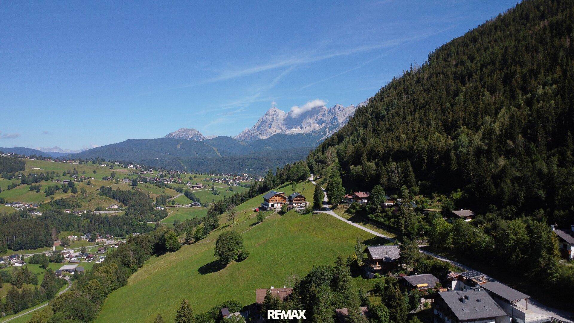 Weitläufige Luftaufnahme einer alpinen Landschaft mit verstreuten Häusern und majestätischen Bergen im Hintergrund.