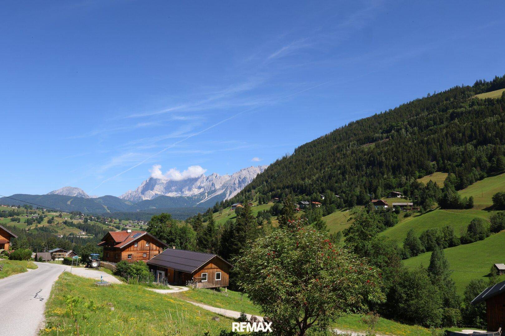 Malerisches Dorf in einem grünen Tal, umgeben von bewaldeten Hügeln und fernen Berggipfeln.