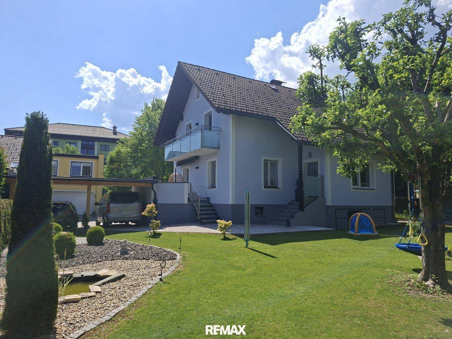 Haus mit hellblauer Fassade, großem Garten, kleinem Teich und Carport an einem sonnigen Tag.