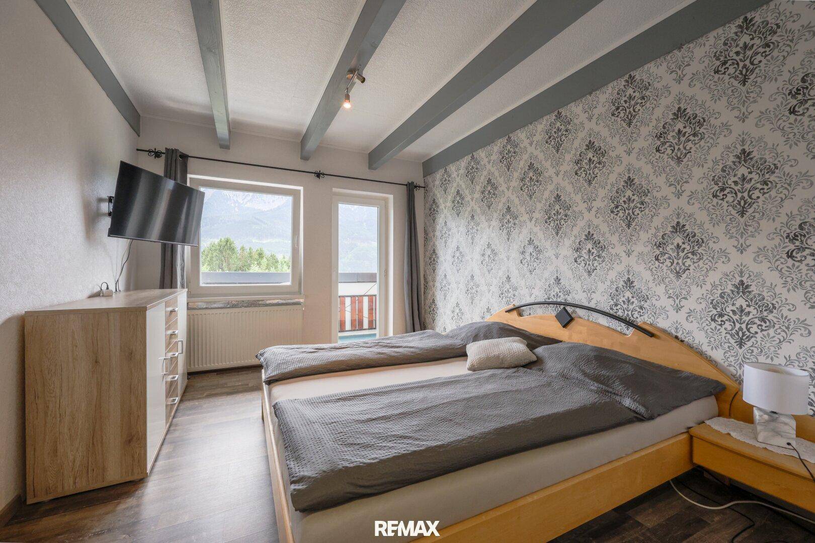 Gemütliches Schlafzimmer mit Holzbalkendecke, gemusterter Tapete und Bergblick vom Fenster.