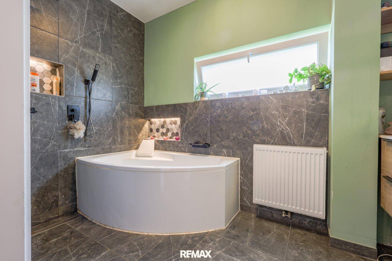 Modernes Badezimmer mit Eckbadewanne, dunklen Fliesen und Fenster für natürliches Licht.