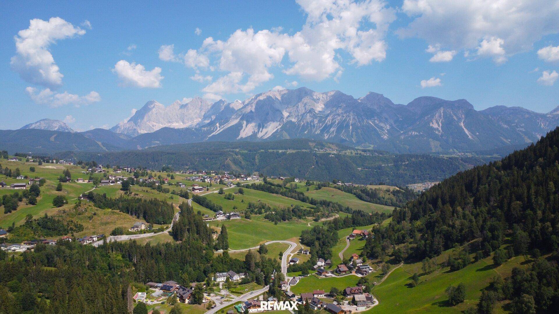 Eine weite Luftaufnahme einer ländlichen Siedlung mit Häusern, Wiesen und Bergen im Hintergrund.