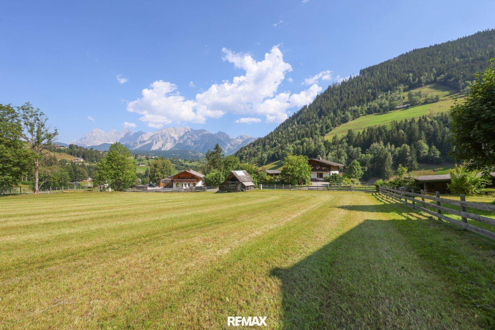 Weitläufige grüne Wiesen und traditionelle Häuser eingebettet in eine malerische Berglandschaft.