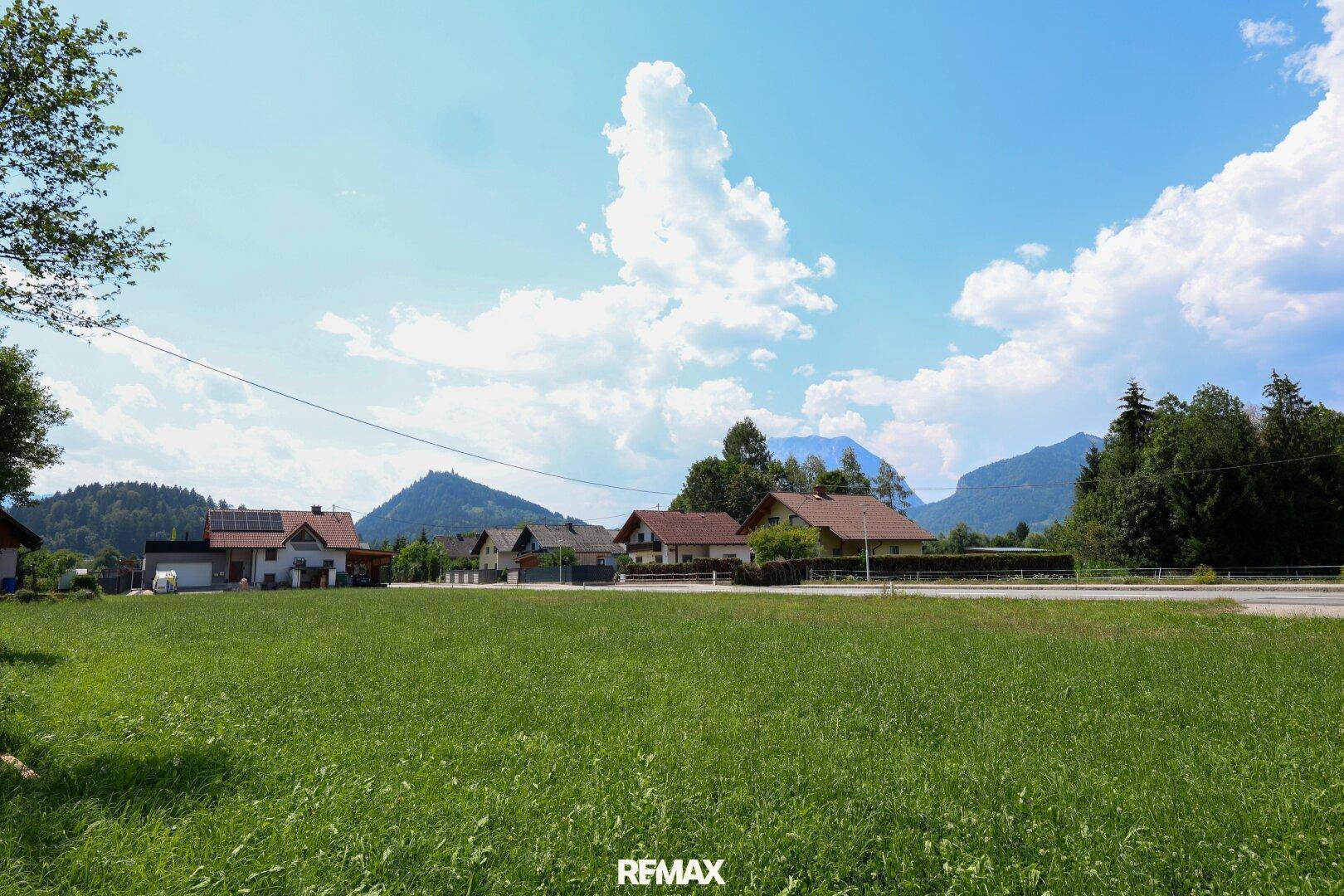 Weitläufige Grünfläche mit Häusern im Hintergrund und einem Panoramablick auf die Berge unter blauem Himmel.