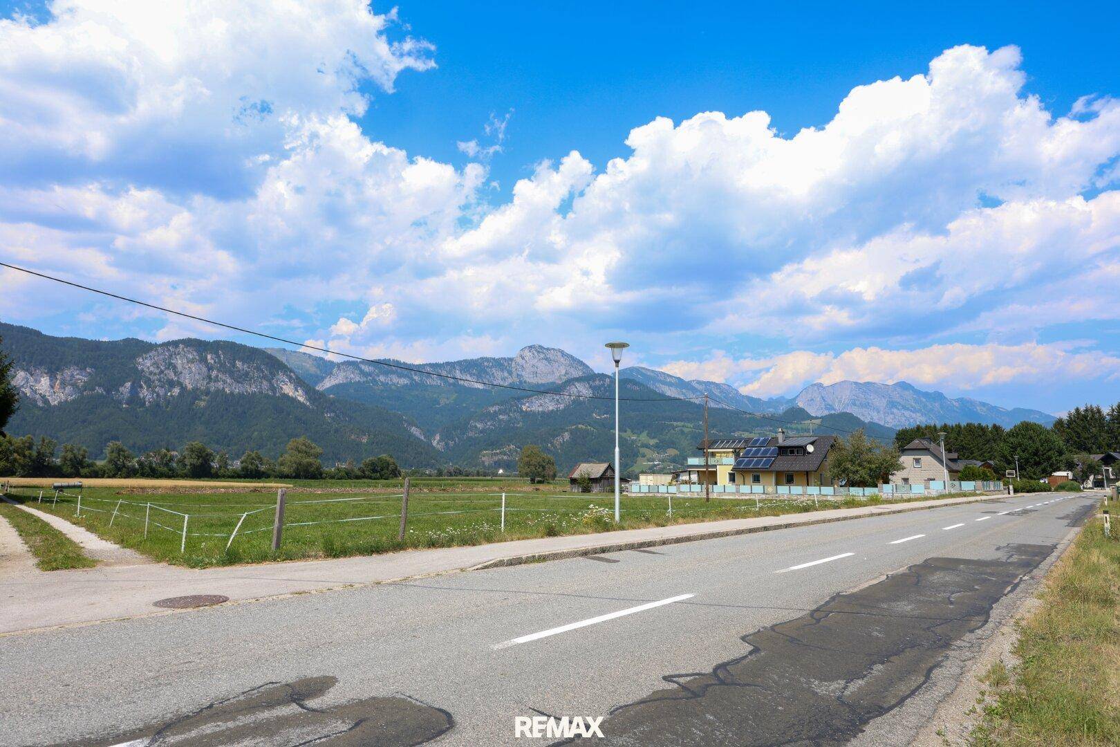 Landstraße führt durch eine ländliche Gegend mit Häusern und beeindruckendem Bergblick unter blauem Himmel.