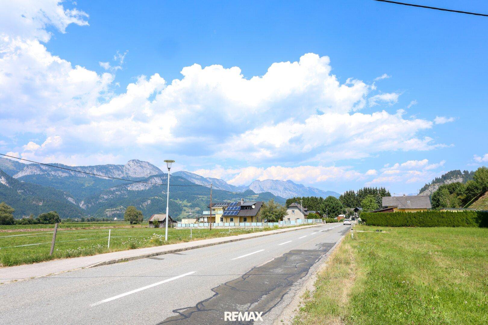 Panoramablick auf eine Landstraße, umgeben von grünen Feldern und einer majestätischen Bergkulisse.