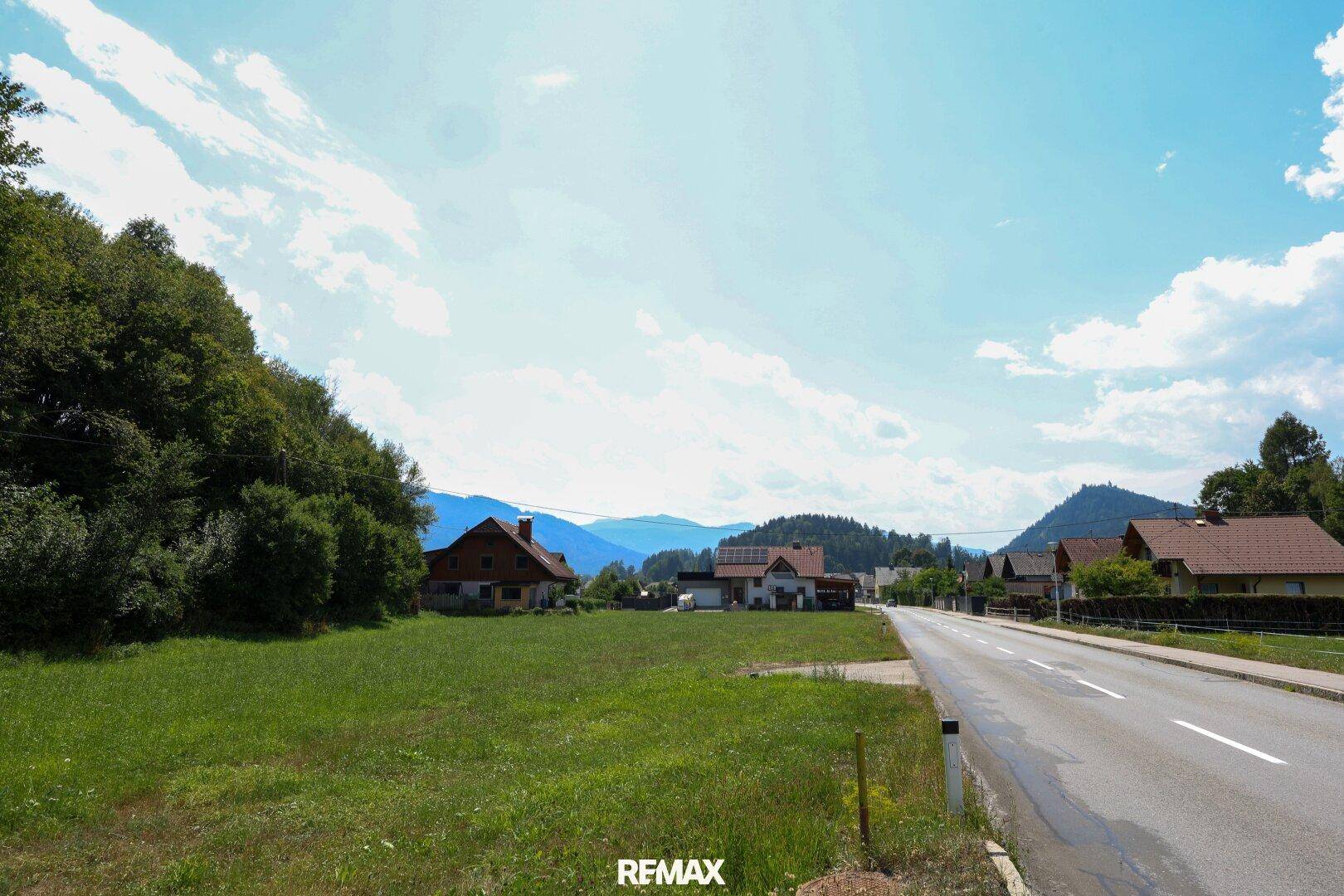 Panoramablick auf ein grünes Grundstück mit angrenzender Straße, Häusern und Bergen im Hintergrund.