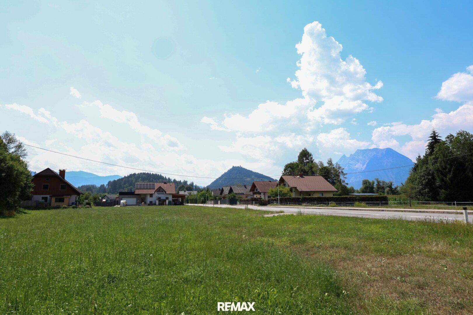Offenes grünes Feld mit Blick auf eine Siedlung, eine Straße und die umliegenden Berge.