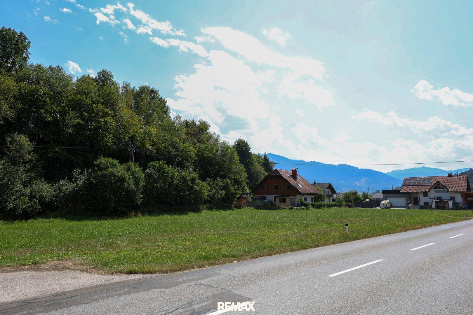 Großes grünes Grundstück mit einem Haus, Bäumen und Bergen im Hintergrund, angrenzend an eine Straße.