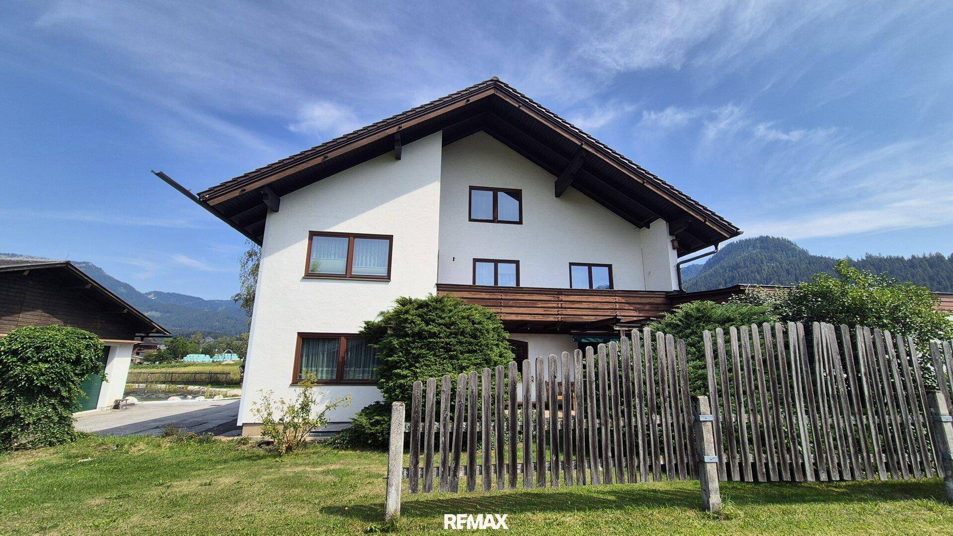 Ansicht des Hauses mit weißer Fassade, braunem Dach und Holzzaun im Vordergrund.