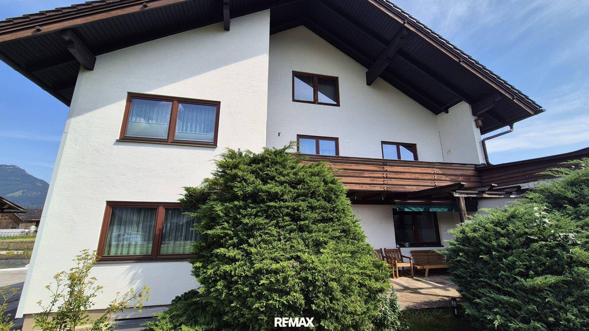 Nahaufnahme der Hausfassade mit weißen Wänden, braunen Fenstern und üppiger Bepflanzung.