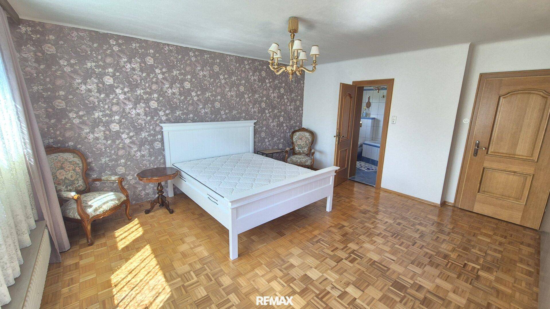 Helles Schlafzimmer mit weißem Bett, floraler Tapete und traditionellen Sitzgelegenheiten.