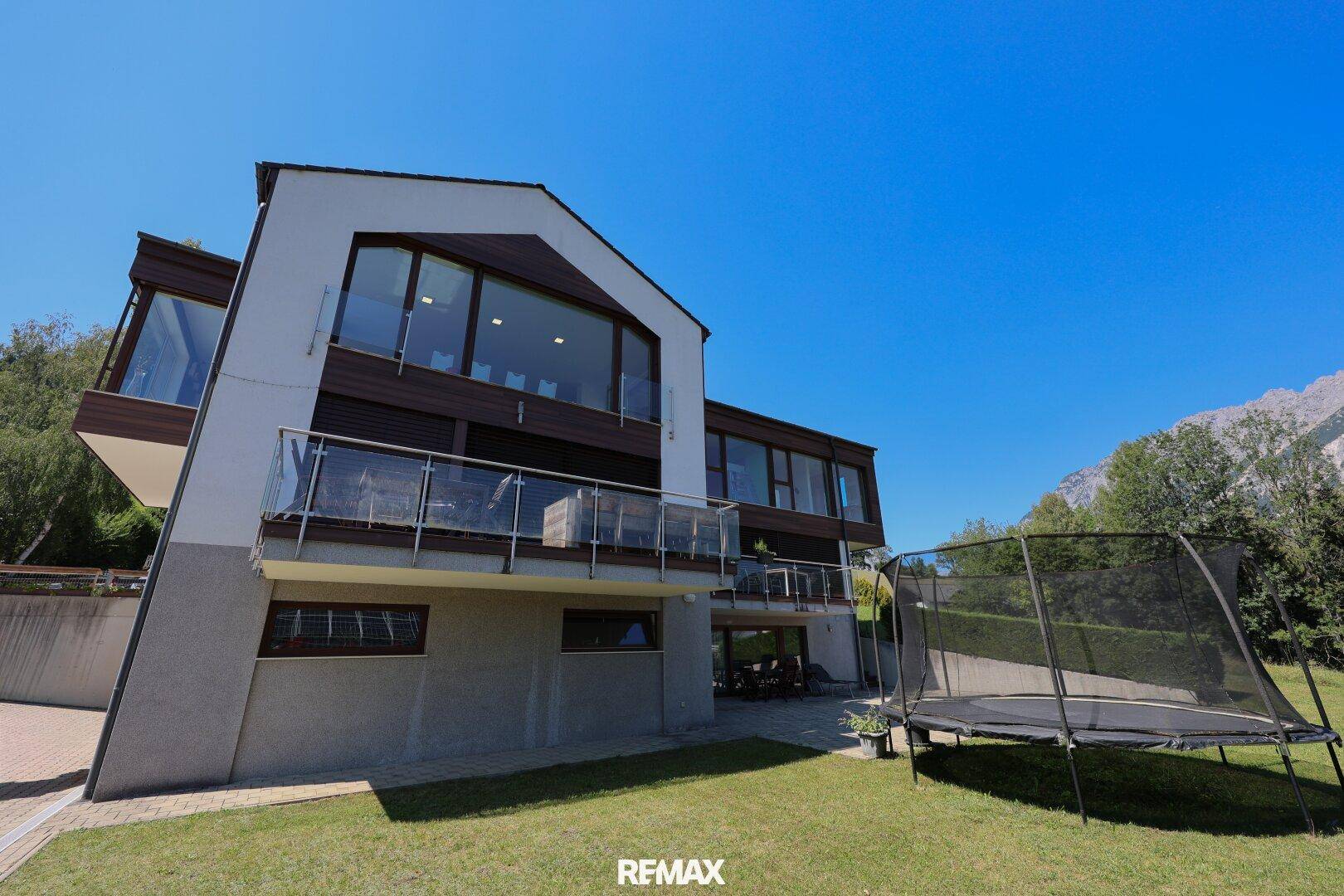 Gartenbereich mit Trampolin und moderner Hausfassade vor Bergkulisse.
