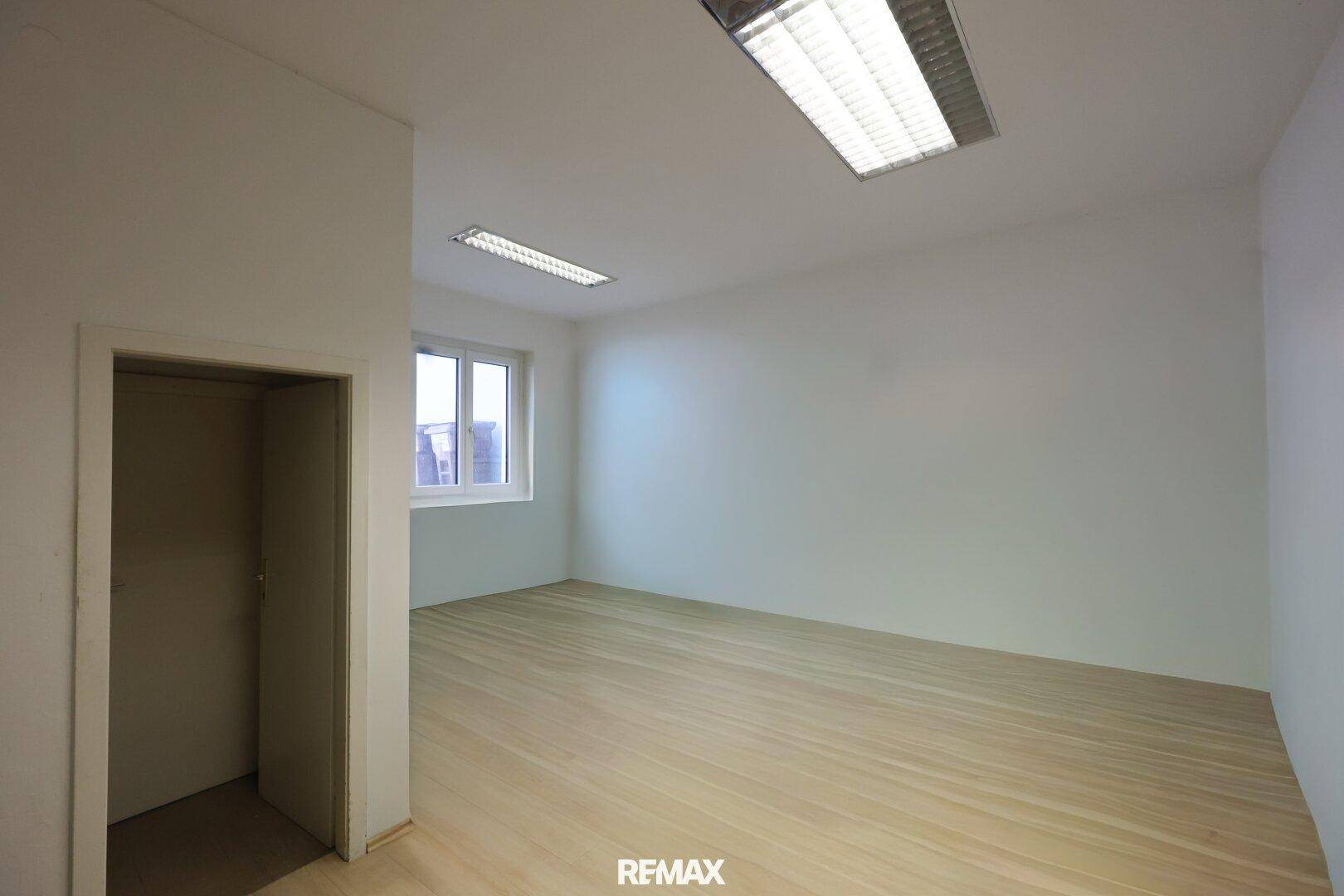 Kleinerer Büroraum mit hellem Holzfußboden, weißen Wänden und einem Fenster für Tageslicht.
