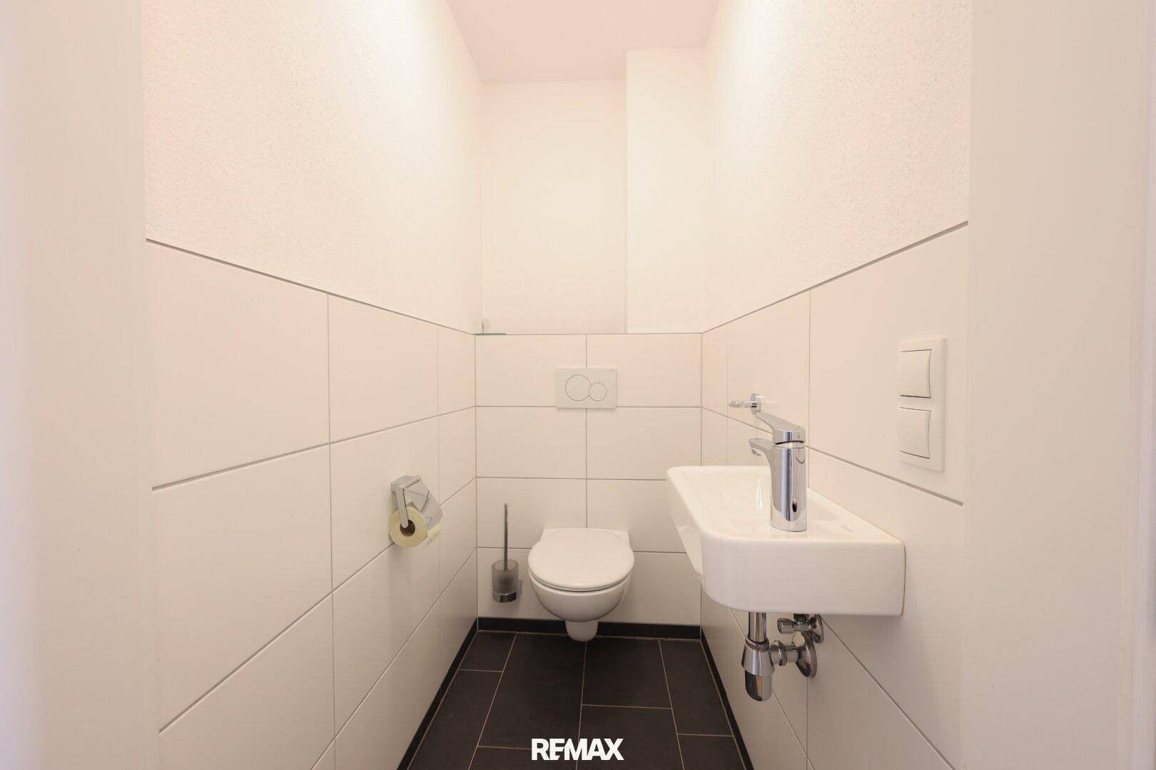 Separates Gäste-WC mit Wand-WC, kleinem Waschbecken und dunklen Bodenfliesen.