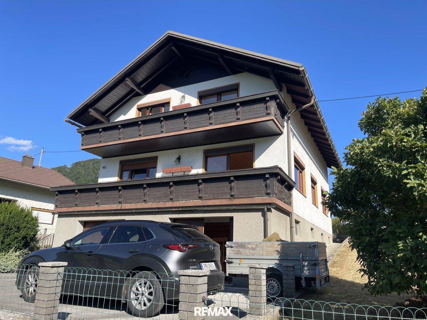 Traditionelles Wohnhaus mit mehreren Balkonen, Satteldach und Parkplatz im Freien vor dem Gebäude.