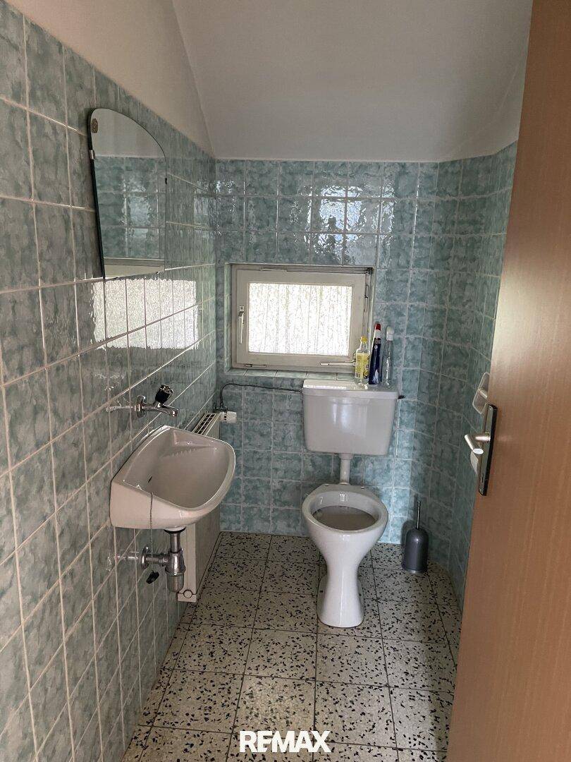 Kleines Badezimmer mit hellblauen Fliesen, WC, Waschbecken und einem Fenster.
