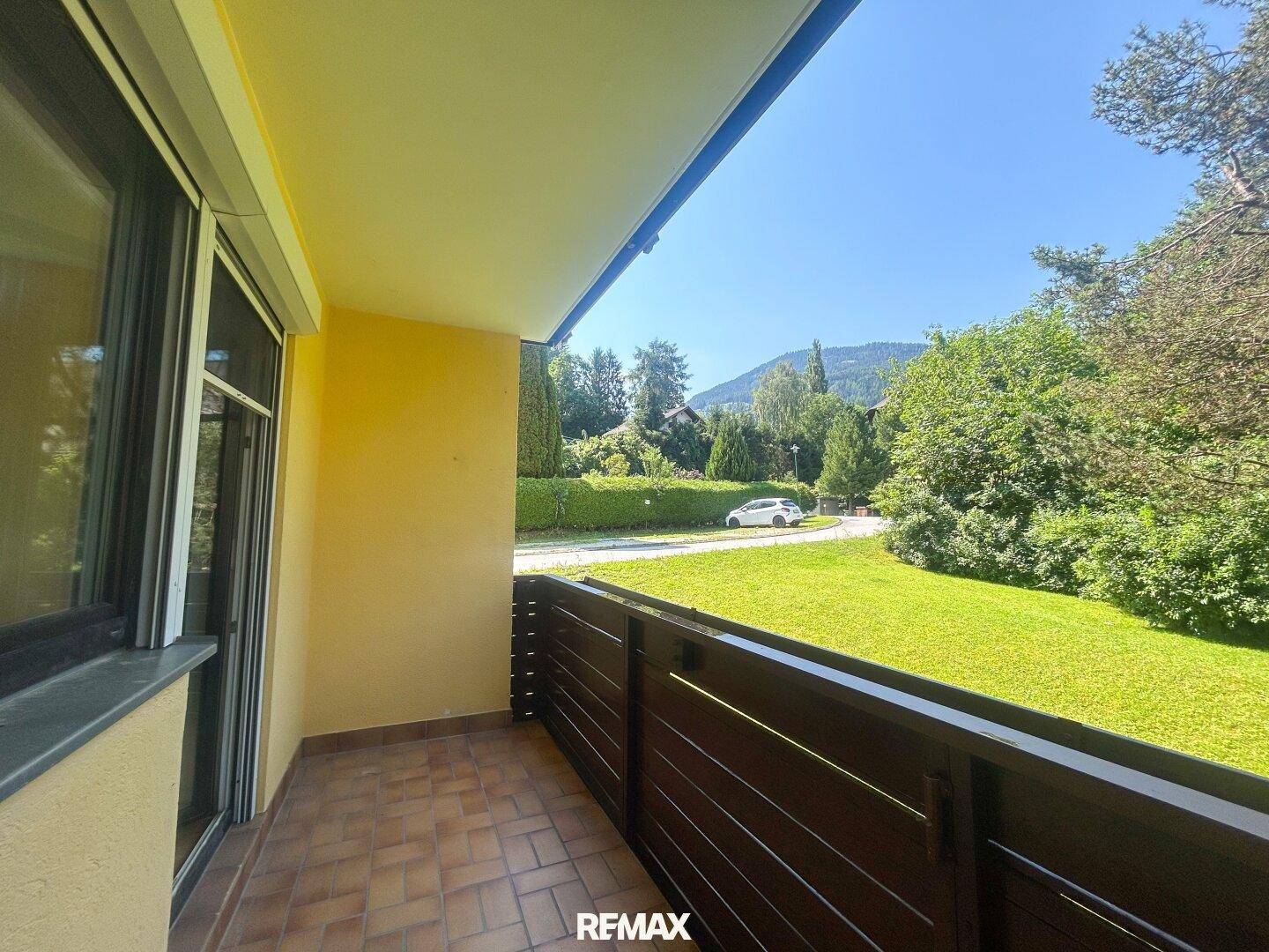 Sonniger Balkon mit Fliesenboden und Holzbalkonbrüstung, bietet einen Blick ins Grüne und auf Berge.