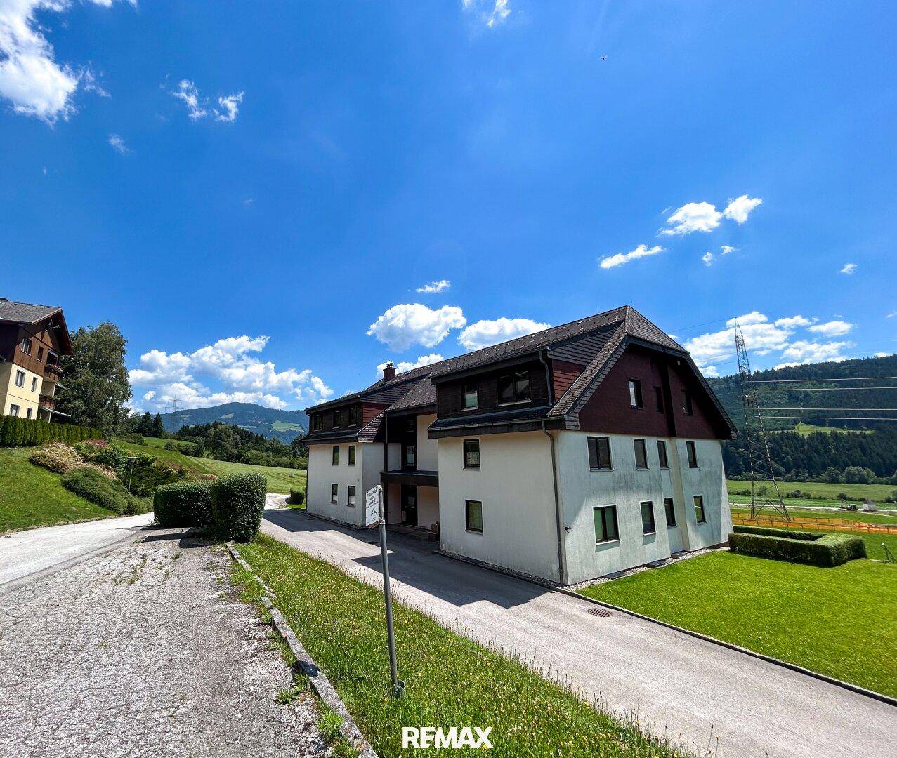 Seitenansicht des Mehrfamilienhauses mit Zufahrt und grüner Umgebung unter strahlend blauem Himmel.